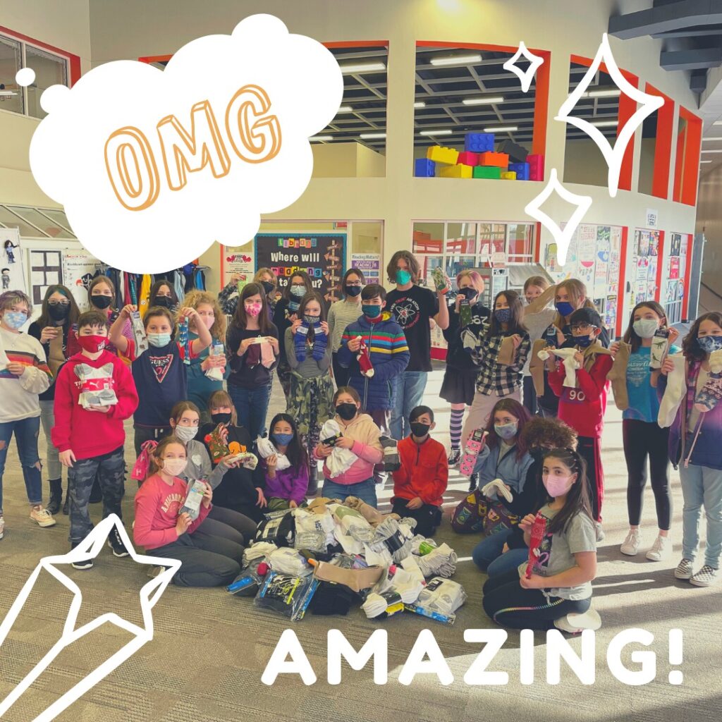 A large group of kids wearing masks pose together indoors, holding items and standing behind a pile of collected goods. Decorative text and graphics say OMG and AMAZING! with stars drawn around the group.
