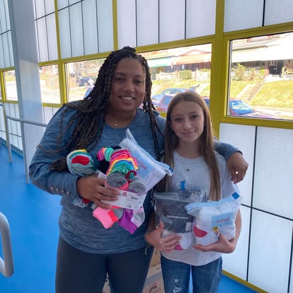 Two people stand indoors, smiling at the camera. The adult holds colorful rolled socks, while the child holds a bag with various items. Large windows behind them show a street and buildings outside.