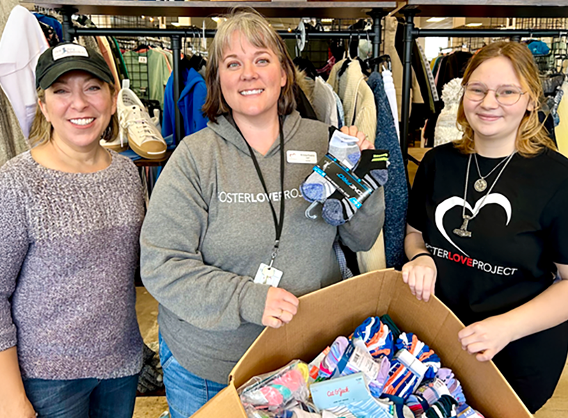 Three women smile while holding and displaying donated socks in a clothing store setting. One woman wears a Foster Love Project hoodie, another a matching t-shirt, and the third a light sweater and cap.