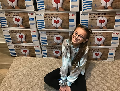 A smiling girl with glasses sits on a carpet in front of a stack of boxes labeled “Have a Heart Book Drive,” each featuring an image of a book with a red heart on the cover.