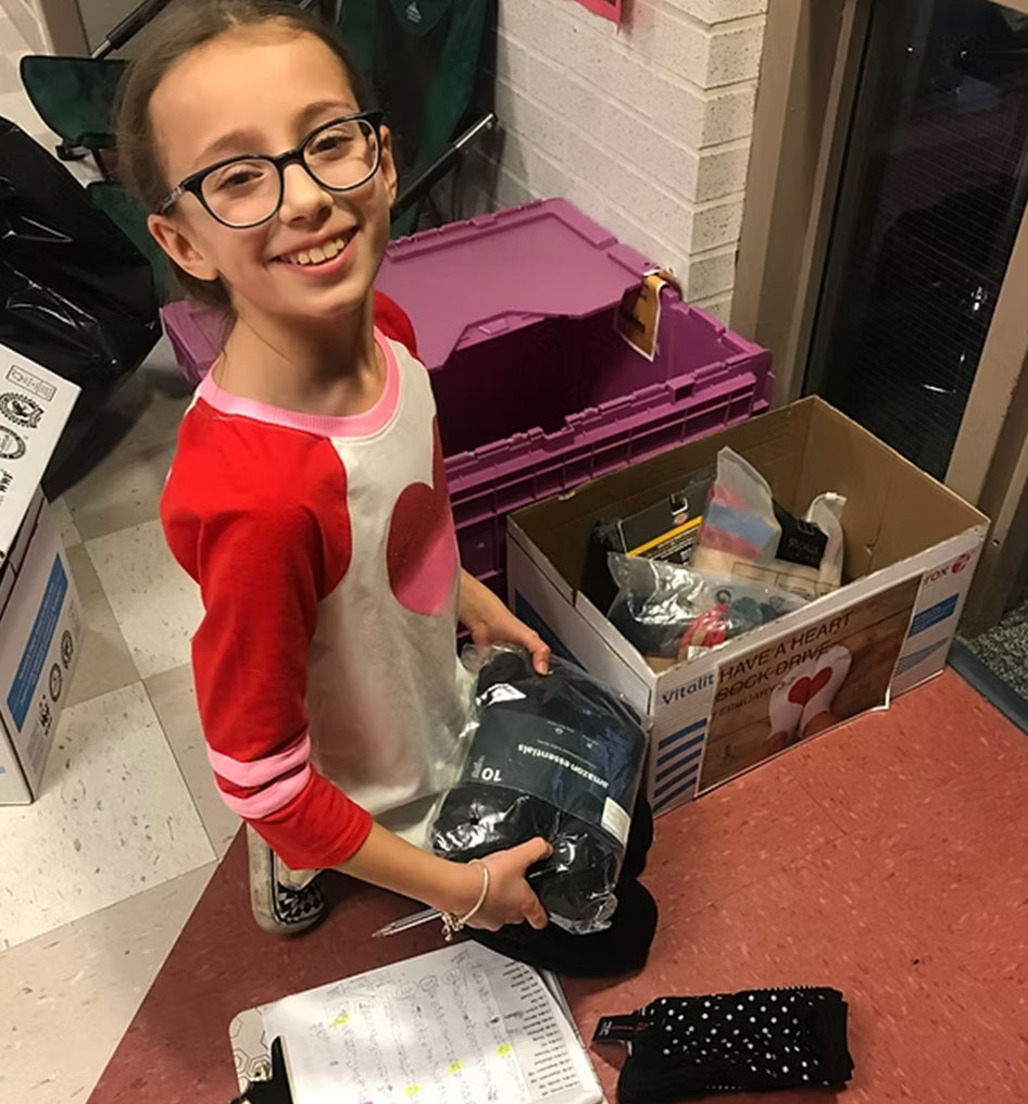 A smiling young girl with glasses kneels on the floor, holding a package. She is next to a box labeled “Have a Heart Sock Drive,” a checklist, and a purple storage bin in a school or community center setting.