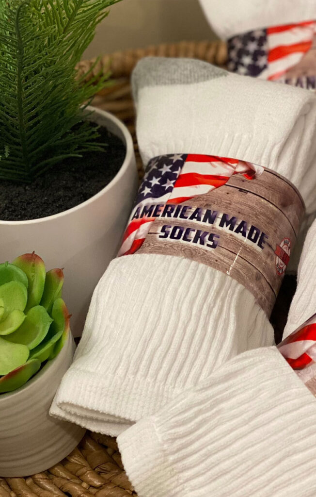 A bundle of white socks labeled American Made Socks with an American flag design on the packaging, placed next to potted plants on a woven surface.