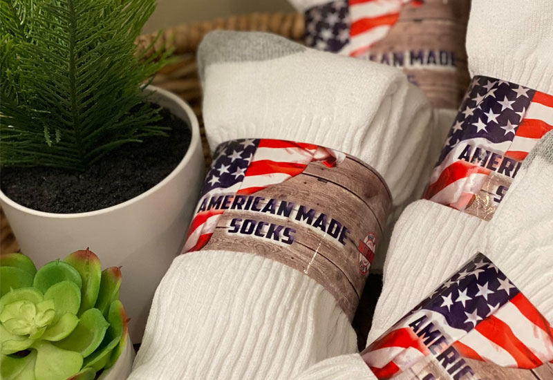 A close-up of several pairs of white socks with labels featuring an American flag and the text American Made Socks, next to a white pot with a small green plant and a succulent.