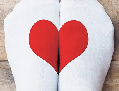 Two feet wearing white socks are placed together side by side, each sock has half of a red heart printed on it, forming a complete heart when the feet are pressed together. The background is wooden.